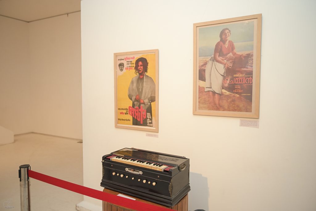 An exhibition space featuring a Pakrashi Harmonium in the foreground, with two framed movie posters on the wall behind it.