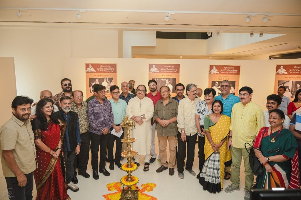 A group of people gathered for the centenary celebration of Salil Chowdhury at the Kolkata Centre for Creativity, featuring a decorative lamp and exhibition banners in the background.